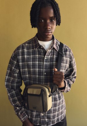 Borsa a tracolla beige con chiusura a zip, caratterizzata da una finitura texture, tenuta da una persona che indossa una camicia a quadri su uno sfondo neutro.
