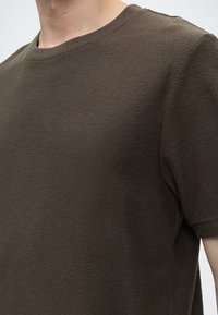 Κοντινό πλάνο ενός ατόμου που φοράει μια υφασμάτινη ελιάς πράσινη κοντομάνικη μπλούζα με στρογγυλή λαιμόκοψη, εμφανίζοντας τον πάνω κορμό και την περιοχή του λαιμού.