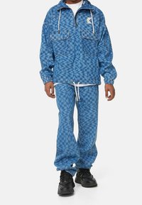 Ensemble en denim bleu composé d'une veste zippée avec deux poches poitrine et d'un pantalon à motifs. Comprend des cordons de serrage et des poignets élastiques.