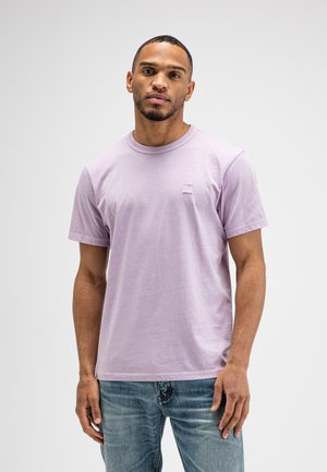 Homme adulte portant un t-shirt violet clair à manches courtes et un jean bleu, debout devant un fond clair uni.