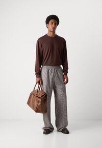 Chemise marron à manches longues assortie à un pantalon large à motifs. Un sac de voyage en cuir marron et des chaussures foncées complètent la tenue.