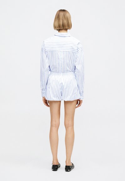 Γυναίκα με κοντά ξανθά μαλλιά στέκεται με την πλάτη, φοράει λευκό μακρυμάνικο πουκάμισο και ταιριαστά ριγέ σορτς με μαύρα παπούτσια.