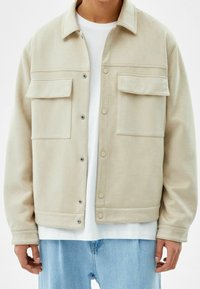 Beige jacket with a smooth texture, button closure, two chest pockets, and a classic collar, worn over a white t-shirt and light blue jeans.