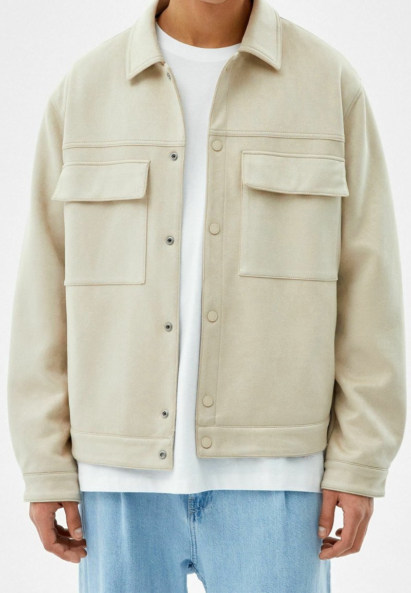 Beige jacket with a smooth texture, button closure, two chest pockets, and a classic collar, worn over a white t-shirt and light blue jeans.