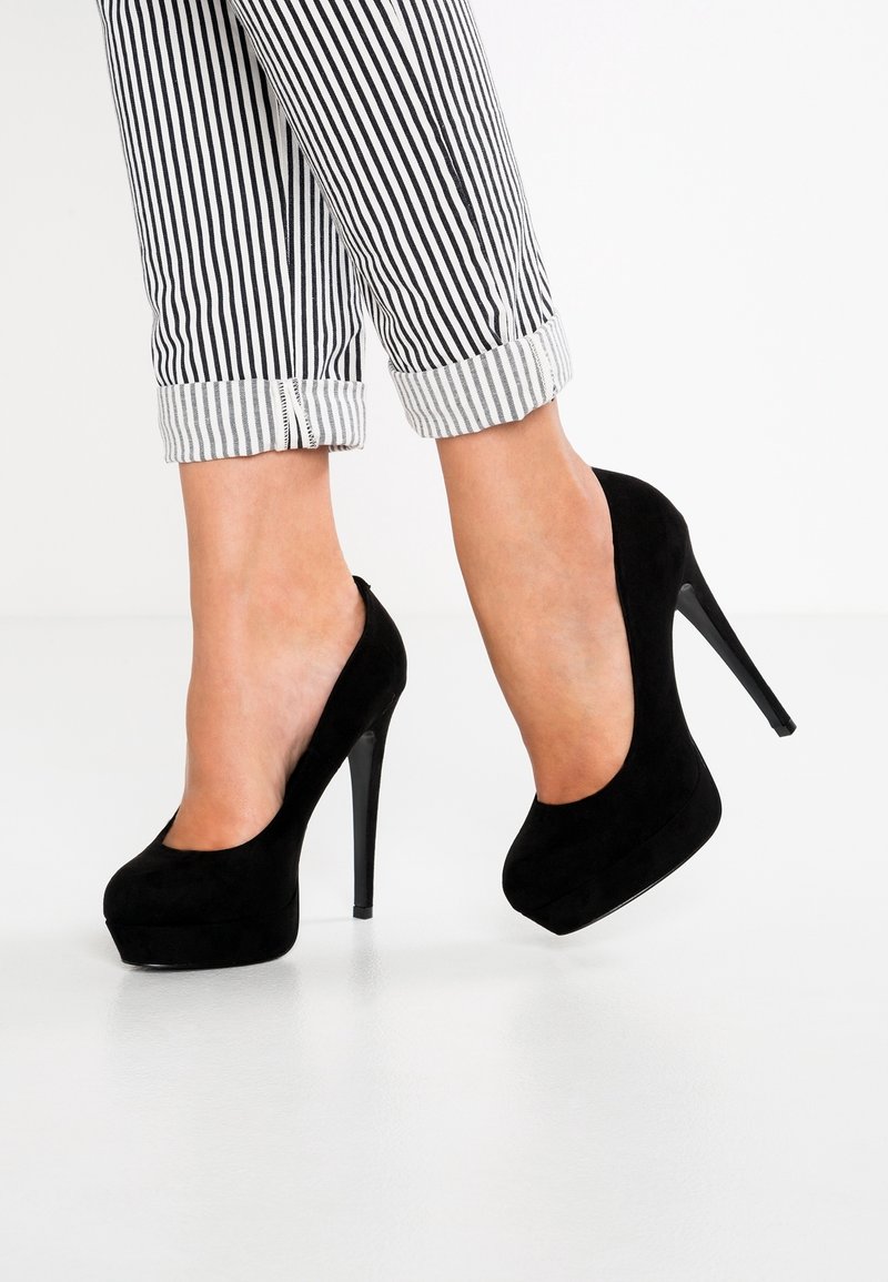Escarpins à talons hauts en daim noir avec une plateforme à l'avant, un bout arrondi et un talon aiguille élancé, assortis à un pantalon rayé noir et blanc retroussé aux chevilles.