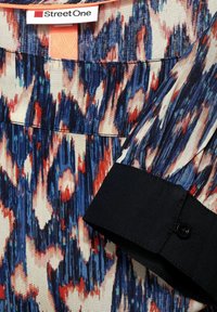 Blusa multicolore con un tessuto a fantasia che presenta disegni astratti blu, rossi e bianchi. Include un polsino nero e un'etichetta con la scritta "Street One."
