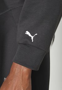 Sweatshirt noir avec une texture lisse, présenté avec un logo Puma blanc sur le poignet. Le design inclut des détails de couture subtils.