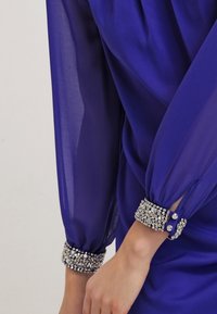 Manga de blusa azul real translúcida con puños anchos decorados con piedras de imitación plateadas y negras, usada en un brazo doblado.