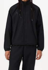 Chaqueta negra con cierre y cuello tipo solapa, puños elásticos y costuras sutiles. El material parece ligero y texturizado.