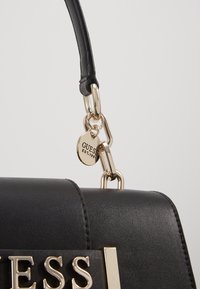 Sac à main synthétique noir à finition brillante, doté de ferrures dorées, d'un accent en maillon de chaîne et d'une étiquette logo affichant "GUESS EST. 1981."