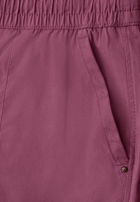 Gros plan sur un pantalon en tissu mauve montrant une taille élastique, une poche latérale avec couture visible, et un petit détail métallique en forme de cœur.