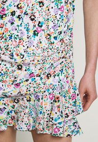 Jupe blanche avec un petit imprimé floral multicolore, ourlet volanté et détails froncés sur le côté, avec une main reposant à côté.