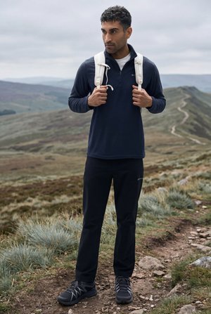 Jaunas vyras, vilkintis tamsius lauko drabužius ir baltą kuprinę, stovi ant akmenuoto žygių tako kalnuotame kraštovaizdyje.