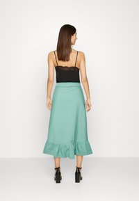 Saia midi verde menta com barra em renda, combinada com um top preto com detalhes em renda. Modelo vista de costas, usando sapatos de salto grossos pretos.