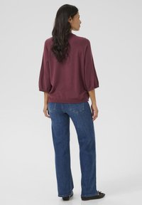 Maglione borgogna con maniche a tre quarti, abbinato a jeans dritti blu e scarpe nere. Il tessuto sembra morbido e testurizzato.