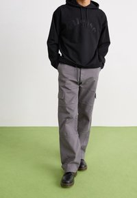 Hombre que lleva una sudadera negra con capucha, pantalones cargo grises con bolsillos laterales y botas negras, de pie sobre un suelo verde claro frente a una pared blanca.