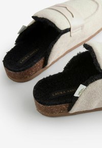 Filzschlappen in Creme und Schwarz, mit einer Korksohle, flauschigem Innenraum und einem glatten Riemen. Logo auf der Innensohle sichtbar.