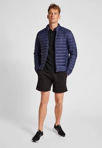 Marinblå vadderad jacka med quiltad design, dragkedja framtill och sidofickor, bärs över en svart pikétröja och svarta shorts, med svarta sportskor.