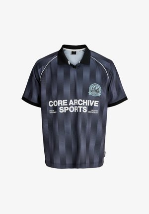 Smiješeći se dječak stoji, obučen u sportsku polo majicu s crnim i sivim prugama preko duge bijele košulje, široke crne hlače i bijele tenisice na jednobojnoj pozadini.