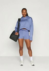 Jeune femme en sweat cropped bleu et shorts assortis, portant des chaussettes blanches et des baskets, portant un sac de sport noir sur une épaule.