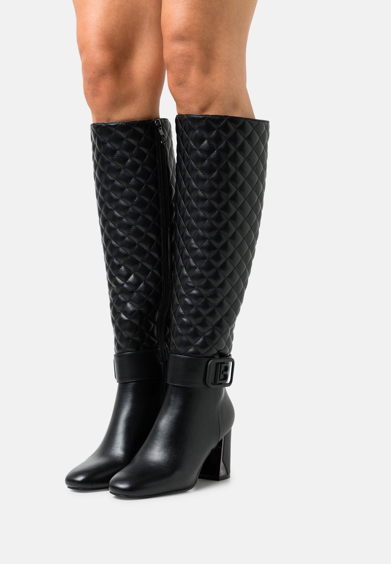 Botas negras acolchadas hasta la rodilla con tacones cuadrados y cremalleras laterales, llevadas sobre piernas desnudas, con un detalle de correa con hebilla por encima del tobillo.