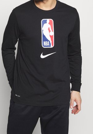 Mężczyzna w czarnej koszulce z długim rękawem z logo NBA i białym znakiem Nike na przodzie, w połączeniu z czarnymi spodniami na jednolitym tle.