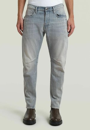 Mann trägt hellgraue Jeans aus Denim und dunkelbraune Lederstiefel und steht vor einem neutralen Hintergrund.