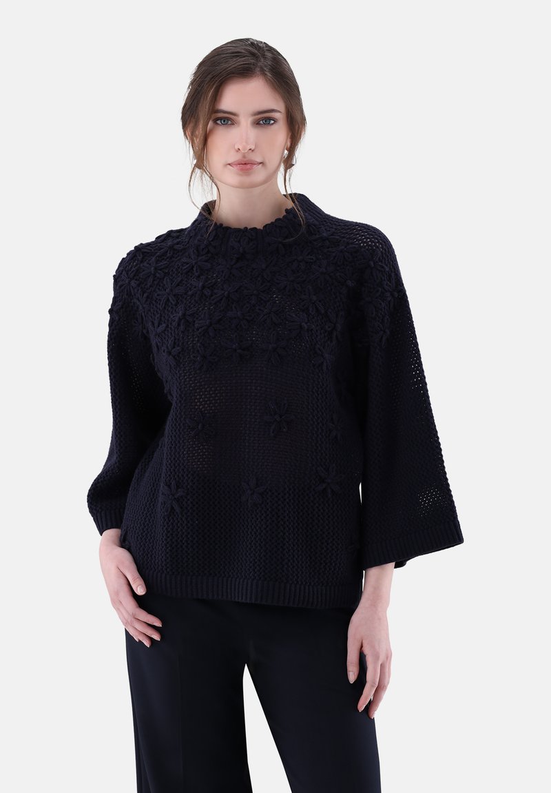 Junge Frau mit lockigem braunem Haar, die einen dunkelblauen Strickpullover mit floralen Mustern und schwarze Hosen trägt, steht vor einem weißen Hintergrund.
