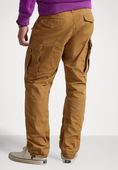 Cargo nohavice z hnedej látky, s viacerými bočnými vreckami, zakriveným zadným sedlom a voľným strihom zúženým ku členkom.