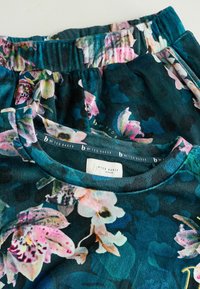 Kvetinová mikina v tmavo tyrkysovej farbe, s jemnou zamatovou textúrou. Dizajn obsahuje ružové a zelené kvetinové vzory. Na štítku je nápis "b by Ted Baker."