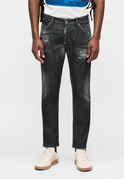 Άνδρας με ξεθωριασμένο στενό τζιν παντελόνι με ελαφρά φθορές και λευκά αθλητικά παπούτσια σε απλό φόντο.