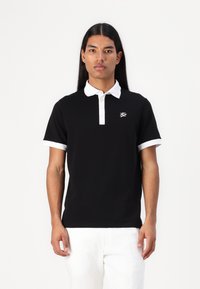 Μαύρο πόλο μπλουζάκι με άσπρο γιακά και μανίκια, με μικρό άσπρο λογότυπο στο στήθος. Το ύφασμα φαίνεται λείο και εφαρμοστό.