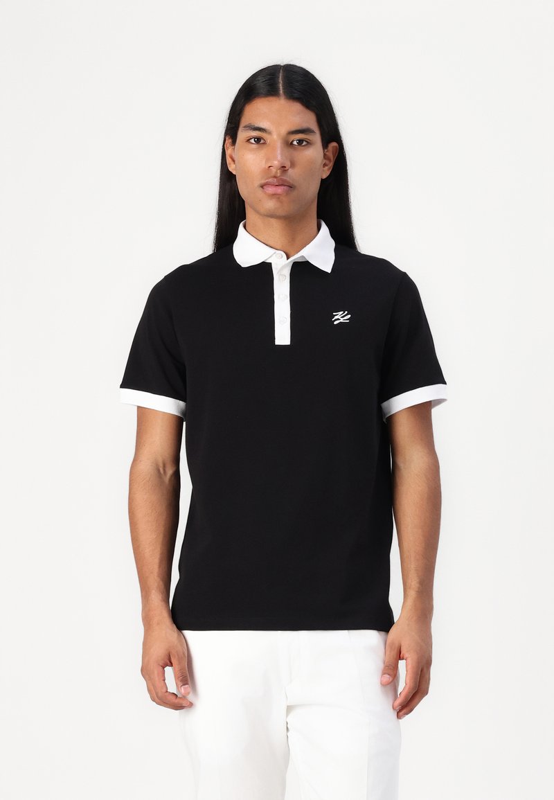 Μαύρο πόλο μπλουζάκι με άσπρο γιακά και μανίκια, με μικρό άσπρο λογότυπο στο στήθος. Το ύφασμα φαίνεται λείο και εφαρμοστό.