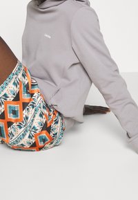 Sudadera con capucha gris y logo blanco, combinada con pantalones cortos de varios colores con un patrón de formas geométricas en naranja, azul y negro.