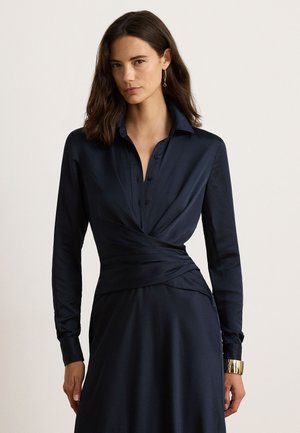 Femme aux cheveux châtain foncé portant une robe bleu marine foncé à manches longues avec un détail portefeuille, un bracelet en or et des boucles d'oreilles pendantes, debout devant un fond uni.