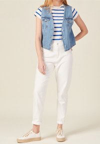 Kamizelka dżinsowa z dwoma przednimi kieszeniami, noszona na białej i niebieskiej, w paski koszulce. Białe spodnie z wysokim stanem i beżowe sneakersy zawiązywane na sznurówki dopełniają stylizację.