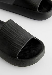 Czarne klapki z teksturowaną górną częścią przypominającą gumę, z otwartym noskiem i płaską, lekko podniesioną podeszwą dla komfortu.