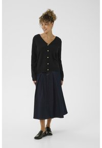 Cardigan nero con scollatura a V e bottoni dorati, abbinato a una gonna di jeans scura di media lunghezza. Il modello indossa mocassini neri, in piedi su uno sfondo bianco.