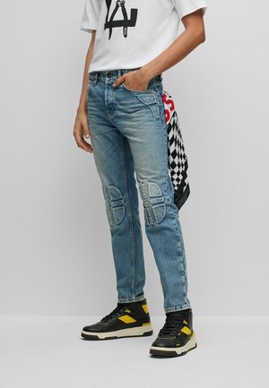 Ljusblå denimjeans med knäfläckar, slim fit och femficksdesign, i kombination med svarta och gula högskor.