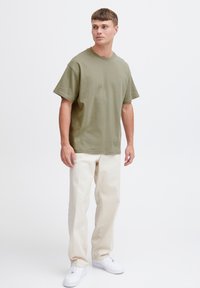 Oliven grøn bomulds t-shirt med rund hals og korte ærmer, parret med beige bukser og hvide sneakers, stående mod en hvid baggrund.