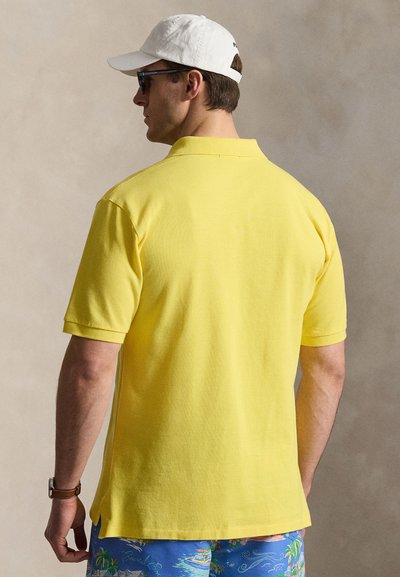 Homme portant un polo jaune manches courtes, un short bleu imprimé tropical, une casquette blanche, des lunettes de soleil et une montre-bracelet marron, debout face à gauche sur fond beige.