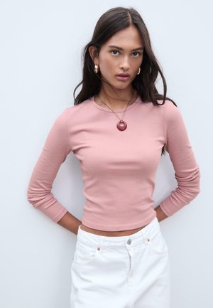 Light pink long-sleeve top with a fitted design. Worn with white pants, accessorized with a large red circular pendant and gold earrings.
