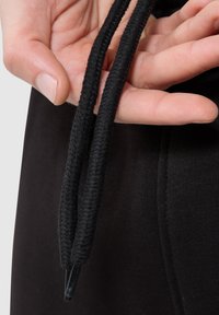 Schwarze Zugbänder in einer Hand, die ein strukturiertes, geflochtenes Material mit Kunststoffspitzen zeigen; befestigt an einem schwarzen Stoffmaterial.