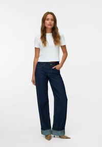 Jeune femme debout avec la main dans la poche, portant un t-shirt blanc court, un jean large sombre avec des ourlets retroussés, et des talons aiguille en imprimé serpent.