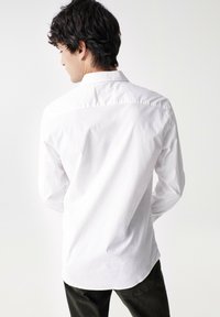 Camisa blanca de manga larga con cuello clásico y detalles de botones, que presenta una textura suave y un dobladillo curvado, combinada con pantalones oscuros.