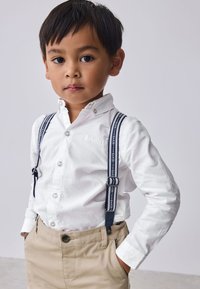White long-sleeve shirt with collar and button closure, paired with beige pants and navy suspenders featuring repeated text. Smooth texture.