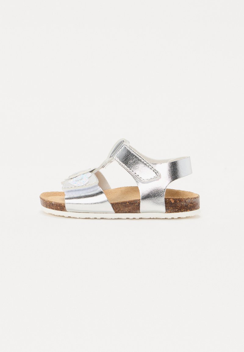 Silver sandals with cork sole, featuring cut-out design and adjustable velcro strap. Accented with a decorative element on the front.