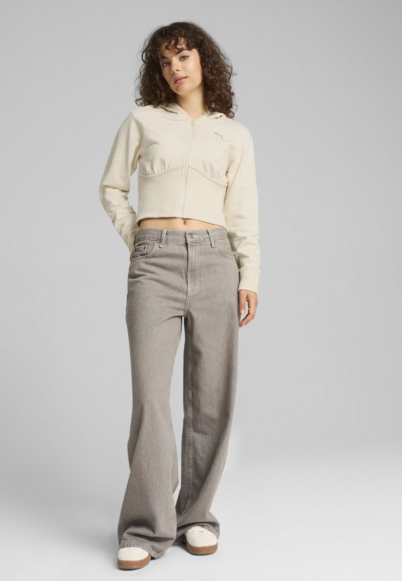 Felpa corta color crema chiaro con zip, abbinata a jeans grigi a gamba larga a vita alta. Scarpe da ginnastica bianche completano l'outfit.