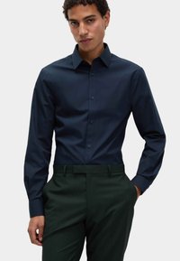 Donker marineblauwe langemouwen overhemd met een button-down kraag, gladde textuur en bijpassende knopen, gedragen met op maat gemaakte donkergroene broek.