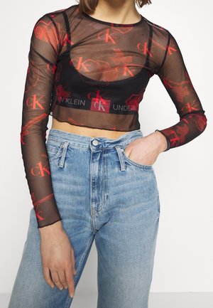 Femme portant un crop top noir semi-transparent à manches longues avec des logos rouges "CK" sur un bralette Calvin Klein noir et un jean taille haute bleu clair.